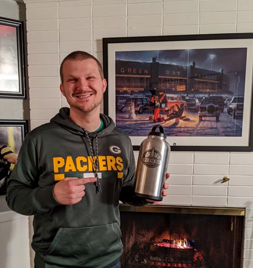 Ben (The Green Bay Guy) holding a water bottle with The Green Bay Guy logo on it in front of a fireplace and Packers memorabilia.